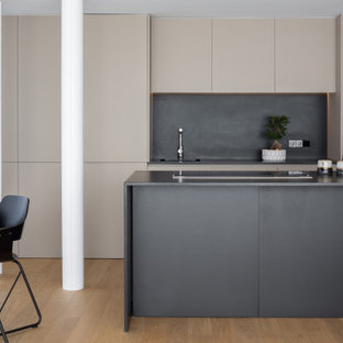 Foto de cocina comedor de galera, actual, de tamaño medio, con armarios con paneles lisos, suelo de madera en tonos medios, una isla, encimeras negras, fregadero integrado, puertas de armario beige, salpicadero negro, electrodomésticos con paneles y suelo marrón