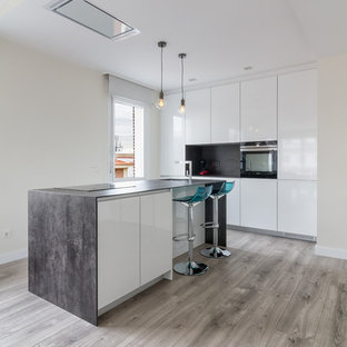 Modelo de cocina lineal, contemporánea, abierta, con armarios con paneles lisos, puertas de armario blancas, salpicadero verde, electrodomésticos negros, suelo de madera en tonos medios, una isla, fregadero bajoencimera, suelo gris y encimeras grises