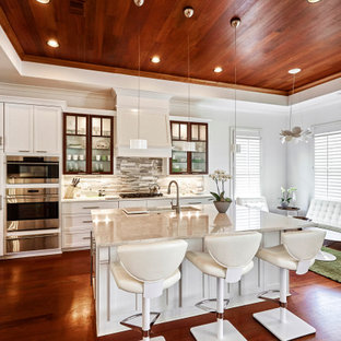 Modelo de cocina en L, tradicional renovada, grande, abierta, con fregadero bajoencimera, armarios con paneles lisos, puertas de armario blancas, encimera de cuarzo compacto, salpicadero beige, salpicadero de azulejos de vidrio, electrodomésticos de acero inoxidable, suelo de madera oscura, una isla, suelo marrón y encimeras beige