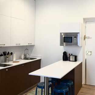 Modelo de cocina actual con armarios con paneles lisos, península, encimeras blancas, fregadero de un seno, puertas de armario blancas, salpicadero blanco, electrodomésticos de acero inoxidable y suelo de madera clara