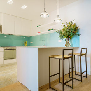Foto de cocina en U, contemporánea, con armarios con paneles lisos, puertas de armario blancas, salpicadero azul, salpicadero de vidrio templado, electrodomésticos de acero inoxidable, suelo de madera clara, península y encimeras grises