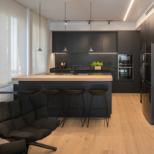 Foto de cocina en L, contemporánea, de tamaño medio, abierta, con puertas de armario negras, fregadero bajoencimera, armarios con paneles lisos, salpicadero negro, electrodomésticos negros, suelo de madera clara, península, suelo beige y encimeras negras