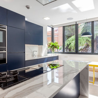 Diseño de cocina comedor contemporánea con fregadero bajoencimera, armarios con paneles lisos, puertas de armario azules, encimera de mármol, salpicadero blanco, salpicadero de mármol, electrodomésticos negros, una isla, suelo beige y encimeras blancas