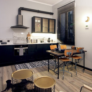 Modelo de cocina lineal, urbana, abierta, sin isla, con puertas de armario negras, salpicadero blanco, electrodomésticos negros, encimeras blancas, armarios estilo shaker, suelo de madera en tonos medios y suelo marrón