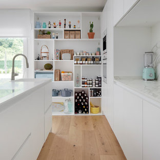 Diseño de cocina lineal, contemporánea, grande, con fregadero integrado, armarios con paneles lisos, puertas de armario blancas, salpicadero blanco, suelo de madera clara, una isla y encimeras blancas