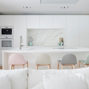 Diseño de cocina lineal, actual, grande, con fregadero integrado, armarios con paneles lisos, puertas de armario blancas, salpicadero blanco, suelo de madera clara, una isla y encimeras blancas