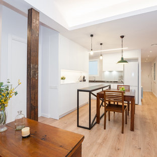 Modelo de cocina actual, grande, abierta, con armarios con paneles lisos, puertas de armario blancas, salpicadero blanco, una isla, encimeras negras, suelo de madera clara y suelo beige