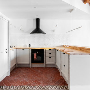 Foto de cocina en U, mediterránea, con fregadero encastrado, armarios estilo shaker, puertas de armario grises, encimera de madera, salpicadero blanco, salpicadero de azulejos tipo metro, electrodomésticos negros, península, suelo rojo y encimeras marrones