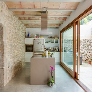 Imagen de cocina lineal, mediterránea, pequeña, abierta, con armarios con paneles lisos, puertas de armario blancas, salpicadero blanco, electrodomésticos de acero inoxidable, suelo de cemento y una isla