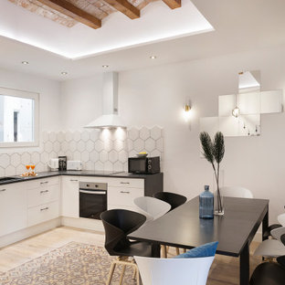 Ejemplo de cocina comedor en L y bandeja, mediterránea, con fregadero encastrado, armarios con paneles lisos, puertas de armario blancas, salpicadero blanco, suelo de madera clara y encimeras negras