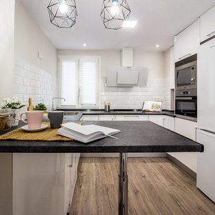 Modelo de cocina en U, contemporánea, pequeña, cerrada, con fregadero encastrado, armarios con paneles lisos, puertas de armario blancas, encimera de laminado, suelo laminado, una isla, encimeras negras, salpicadero blanco, salpicadero de azulejos tipo metro, electrodomésticos blancos y suelo marrón