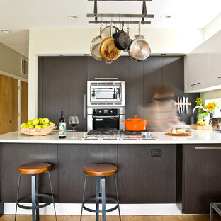 Ejemplo de cocina en U, contemporánea, de tamaño medio, con armarios con paneles lisos, puertas de armario negras, electrodomésticos de acero inoxidable, península, suelo de madera en tonos medios, suelo marrón, encimeras blancas y salpicadero blanco
