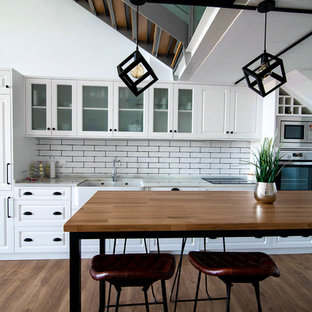 Imagen de cocina lineal, tradicional renovada, con fregadero sobremueble, armarios con paneles con relieve, puertas de armario blancas, salpicadero blanco, salpicadero de azulejos tipo metro, electrodomésticos de acero inoxidable, suelo de madera en tonos medios, una isla, suelo marrón y encimeras blancas