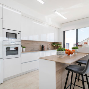 Imagen de cocina de galera, actual, con armarios con paneles lisos, puertas de armario blancas, encimera de madera, salpicadero beige, salpicadero de madera, electrodomésticos blancos, península, suelo beige y encimeras beige