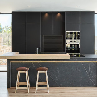 Modelo de cocina moderna con armarios con paneles lisos, puertas de armario negras, electrodomésticos negros, suelo de madera clara, una isla, suelo beige y encimeras grises
