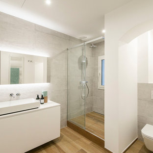 Modelo de cuarto de baño mediterráneo con armarios con paneles lisos, puertas de armario blancas, sanitario de pared, baldosas y/o azulejos grises, paredes blancas, lavabo sobreencimera, suelo marrón, encimeras blancas y ducha con puerta corredera