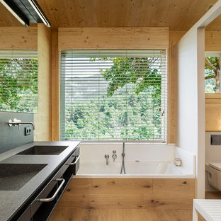 Imagen de cuarto de baño principal, moderno, con armarios con paneles lisos, puertas de armario negras, bañera empotrada, sanitario de pared, paredes blancas, suelo de madera clara, lavabo integrado y encimeras negras