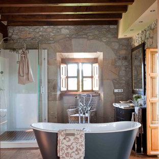 Modelo de cuarto de baño piedra, mediterráneo, con bañera exenta, ducha esquinera, suelo de madera en tonos medios, suelo marrón y ducha con puerta con bisagras