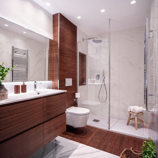 Modelo de cuarto de baño con ducha, contemporáneo, con armarios con paneles lisos, puertas de armario de madera oscura, ducha empotrada, sanitario de pared, paredes blancas, lavabo integrado, suelo multicolor y encimeras blancas