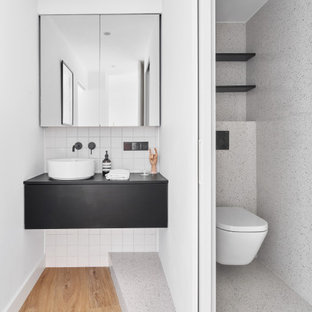 Imagen de cuarto de baño cuarto de baño, único y flotante, minimalista, con armarios con paneles lisos, puertas de armario negras, sanitario de pared, baldosas y/o azulejos blancos, paredes blancas, suelo de madera en tonos medios, lavabo sobreencimera, suelo marrón y encimeras negras