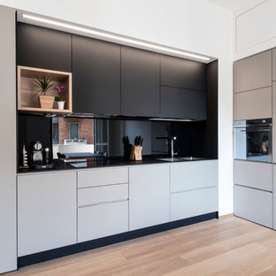 Inspiration for a mid-sized contemporary l-shaped kitchen in Milan with a double-bowl sink, flat-panel cabinets, laminate benchtops, black splashback, glass sheet splashback, stainless steel appliances, light hardwood floors, no island and grey cabinets.