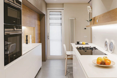 На фото: параллельная кухня в современном стиле с плоскими фасадами, белыми фасадами, серым полом, белой столешницей, белым фартуком и черной техникой без острова с