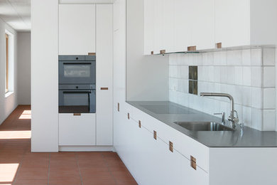 他の地域にある中くらいなインダストリアルスタイルのおしゃれなL型キッチン (シングルシンク、フラットパネル扉のキャビネット、白いキャビネット、ステンレスカウンター、白いキッチンパネル、テラコッタタイルのキッチンパネル、シルバーの調理設備、テラコッタタイルの床、赤い床、グレーのキッチンカウンター) の写真