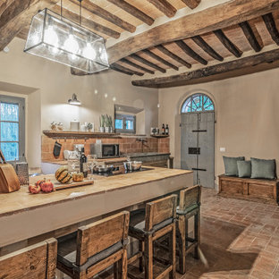 This is an example of a large country galley kitchen/diner in Florence with a belfast sink, beaded cabinets, distressed cabinets, tile countertops, stainless steel appliances, brick flooring, an island, orange floors, orange worktops and exposed beams.