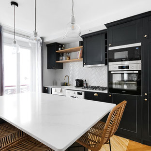 Aménagement d'une cuisine contemporaine avec un évier de ferme, un placard à porte shaker, des portes de placard noires, une crédence blanche, une crédence en carreau de céramique, un électroménager noir, un sol en bois brun, un îlot central, un sol beige et un plan de travail blanc.