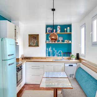Cette photo montre une cuisine américaine éclectique en L de taille moyenne avec un plan de travail en bois, un électroménager de couleur, un sol en carreau de terre cuite, un sol marron, un évier posé, des portes de placard blanches et une crédence bleue.