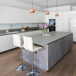 Réalisation d'une grand cuisine ouverte design en L avec un évier 1 bac, des portes de placard blanches, un plan de travail en granite, une crédence blanche, un électroménager noir, un îlot central, un sol marron, un plan de travail gris, un placard à porte plane, une crédence en feuille de verre et un sol en bois foncé.
