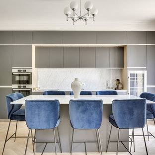 Inspiration pour une grand cuisine américaine linéaire design avec des portes de placard grises, un plan de travail en marbre, une crédence blanche, une crédence en marbre, un électroménager en acier inoxydable, un sol en bois clair, un îlot central, un plan de travail blanc, un évier encastré, un placard à porte affleurante et un sol marron.