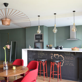 Idées déco pour une cuisine américaine parallèle contemporaine avec un placard à porte plane, des portes de placards vertess, un plan de travail en quartz, un électroménager en acier inoxydable, un sol en bois clair, un sol beige, un plan de travail blanc et une péninsule.
