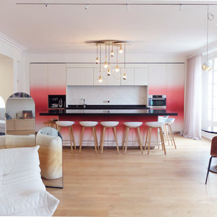 Réalisation d'une grand cuisine ouverte linéaire design avec un évier encastré, des portes de placard rouges, un plan de travail en granite, une crédence blanche, une crédence en marbre, un électroménager en acier inoxydable, un sol en bois clair, un îlot central, un plan de travail noir, un placard à porte plane et un sol beige.