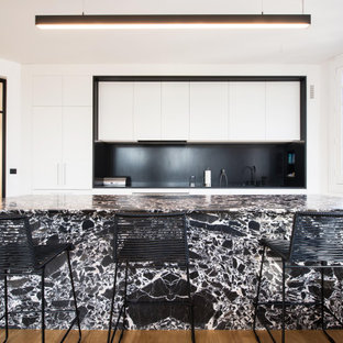 Idées déco pour une grand cuisine parallèle contemporaine avec un plan de travail en granite, une crédence noire, un sol en bois brun, un îlot central, un plan de travail noir, un placard à porte plane, des portes de placard blanches, une crédence en dalle de pierre, un électroménager encastrable et un sol marron.