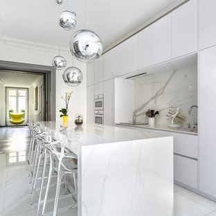 Aménagement d'une cuisine parallèle contemporaine fermée avec un placard à porte plane, des portes de placard blanches, une crédence blanche, une crédence en dalle de pierre, un îlot central et un sol blanc.