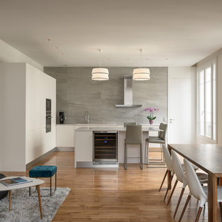 Cette photo montre une cuisine ouverte tendance en L de taille moyenne avec un évier encastré, un placard à porte plane, des portes de placard blanches, une crédence grise, un îlot central, une crédence en carrelage de pierre, un électroménager noir, un plan de travail blanc, un sol en bois clair et un sol marron.