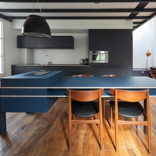 Cette image montre une cuisine ouverte linéaire nordique avec un placard à porte plane, des portes de placard noires, un électroménager en acier inoxydable, un sol en bois brun, un îlot central, un évier encastré, un plan de travail bleu, une crédence blanche et un sol marron.