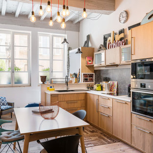 Exemple d'une cuisine américaine éclectique en L avec un évier posé, un placard à porte plane, des portes de placard en bois brun, une crédence noire, un électroménager noir, un sol en bois clair, aucun îlot et un sol beige.