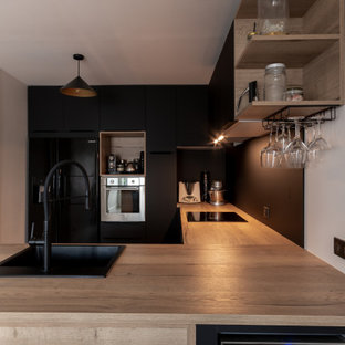 Cette photo montre une cuisine tendance en U de taille moyenne avec un évier posé, un placard à porte plane, des portes de placard noires, un plan de travail en bois, une crédence noire, une crédence en carreau de porcelaine, un électroménager noir et un plan de travail beige.