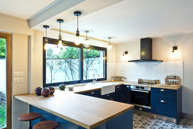 Large retro u-shaped open plan kitchen in Paris with a belfast sink, recessed-panel cabinets, blue cabinets, wood worktops, white splashback, metro tiled splashback, stainless steel appliances, cement flooring, an island, multi-coloured floors and multicoloured worktops.