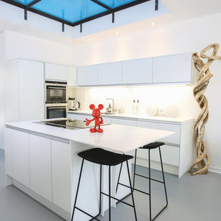 Cette photo montre une cuisine tendance en L de taille moyenne avec un évier 1 bac, des portes de placard blanches, un plan de travail en béton, une crédence blanche, une crédence en dalle de pierre, un électroménager encastrable, béton au sol, un îlot central, un sol gris, un plan de travail blanc et un placard à porte plane.