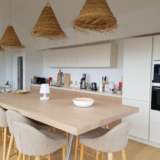 Cette photo montre une grand cuisine américaine parallèle bord de mer avec un évier encastré, un placard à porte affleurante, des portes de placard beiges, un plan de travail en quartz, un sol en bois clair, un îlot central, une crédence blanche, un électroménager noir et un sol beige.
