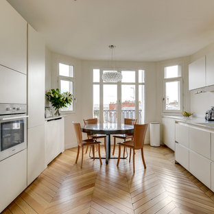 Aménagement d'une grand cuisine américaine parallèle contemporaine avec un évier intégré, une crédence blanche, un électroménager encastrable, un sol en bois clair, un plan de travail blanc, un placard à porte plane, aucun îlot, un sol beige et des portes de placard blanches.