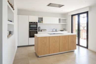 Cette image montre une cuisine design avec un placard à porte plane, des portes de placard blanches, une crédence blanche, un électroménager noir, îlot, un sol beige et un plan de travail blanc.