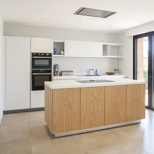 Cette image montre une cuisine design avec un placard à porte plane, des portes de placard blanches, une crédence blanche, un électroménager noir, un îlot central, un sol beige et un plan de travail blanc.