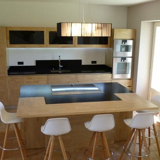 This is an example of a large contemporary u-shaped open plan kitchen in Marseille with an integrated sink, flat-panel cabinets, light wood cabinets, granite benchtops, black splashback, stainless steel appliances, terra-cotta floors, with island, blue floor and black benchtop.