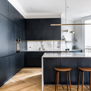 Exemple d'une cuisine tendance en L avec un évier encastré, un placard à porte plane, des portes de placard noires, une crédence blanche, un électroménager noir, un sol en bois brun, un îlot central, un sol marron et un plan de travail blanc.