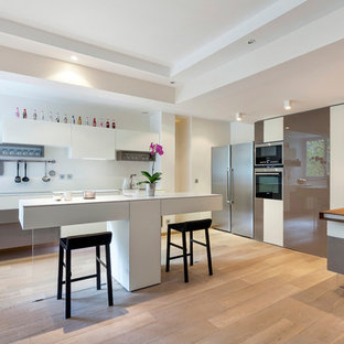 Idée de décoration pour une grand cuisine américaine design en L avec un évier encastré, un placard à porte vitrée, des portes de placard blanches, un plan de travail en quartz, une crédence blanche, une crédence en carreau de verre, un électroménager en acier inoxydable, un sol en bois clair et 2 îlots.