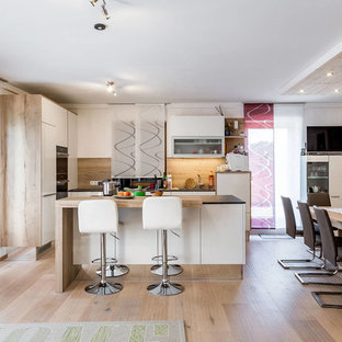Exemple d'une cuisine ouverte tendance en L avec un placard à porte plane, des portes de placard blanches, une crédence beige, une crédence en bois, un électroménager encastrable, un sol en bois clair, un îlot central, un sol beige et un plan de travail noir.